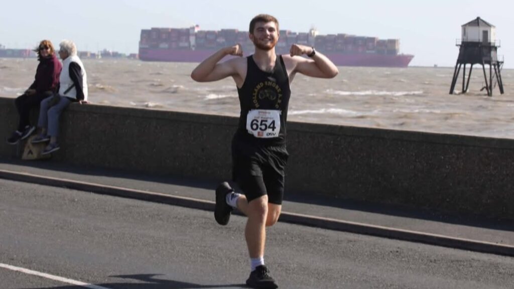 Image shows Alex running in a black running vest and shorts along a coastal road with the sea in the background