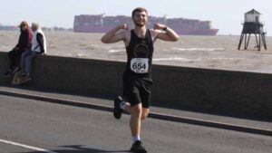 Image shows Alex running in a black running vest and shorts along a coastal road with the sea in the background