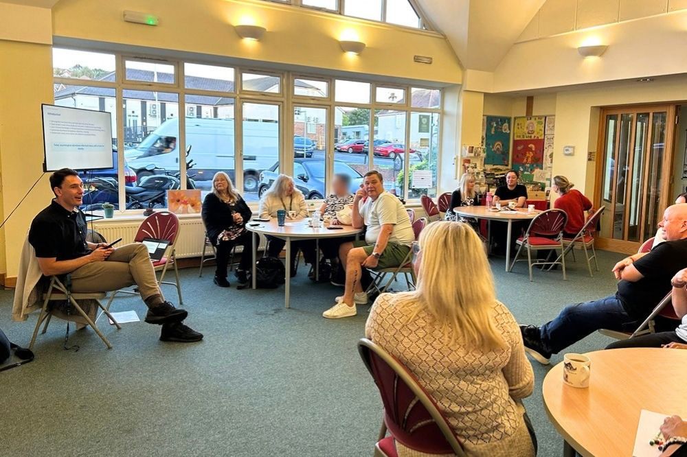 Image shows a room with people sitting at round tables in small groups looking towards a person who is speaking at the front of the room