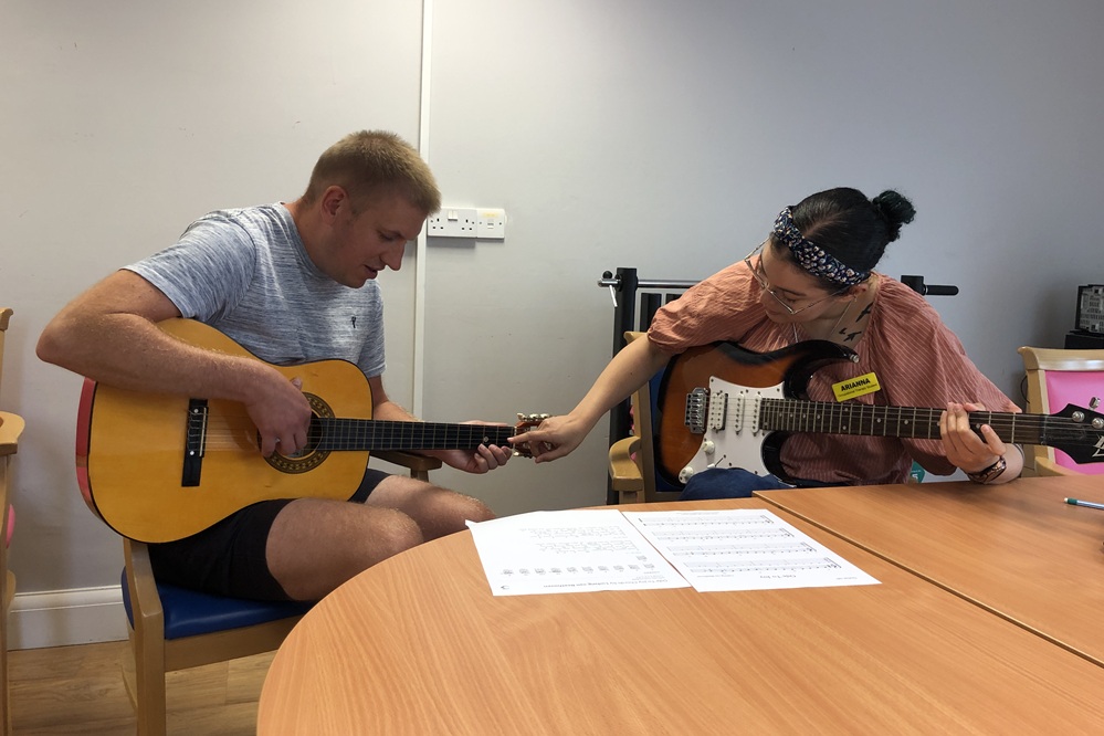 Image shows two people with acoustic guitars. The person on the right is pointing to the fret board on the other person's guitar to guide him.